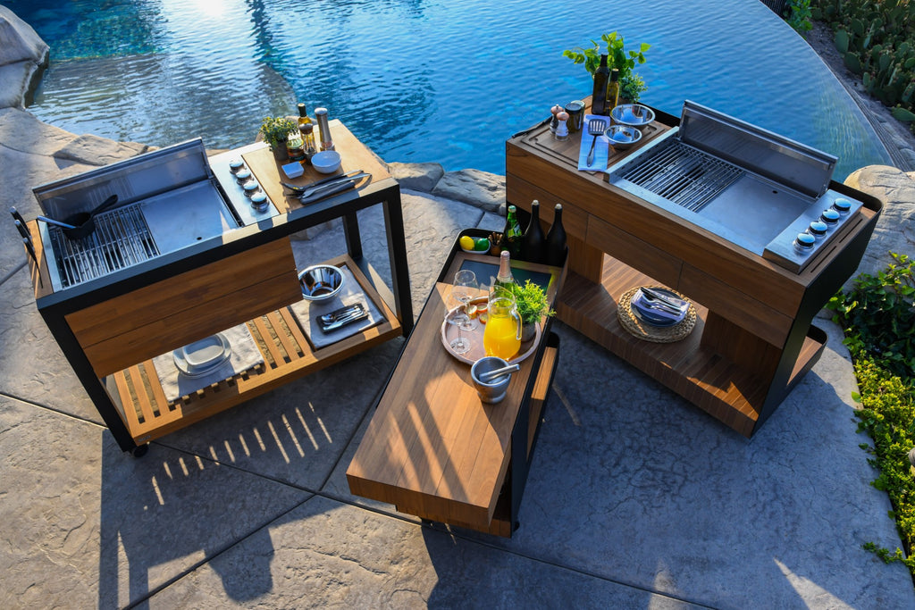 Three luxury indu+ outdoor kitchen carts beside a pool: one with a cooking range, one as a bar, and one for prep, all with dark wood and steel finishes, embodying poolside elegance.