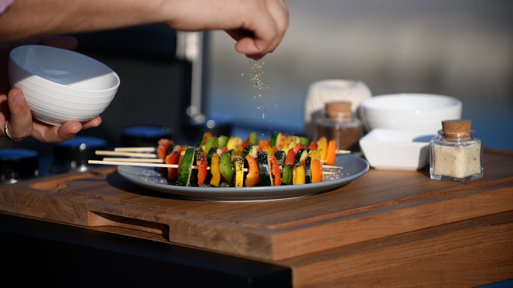 A hand adding a flavorful seasoning to freshly grilled vegetable skewers prepared on the Bistro Island by Indu+. The perfectly cooked skewers are ready to be served, showcasing a mouthwatering combination of colors and flavors.